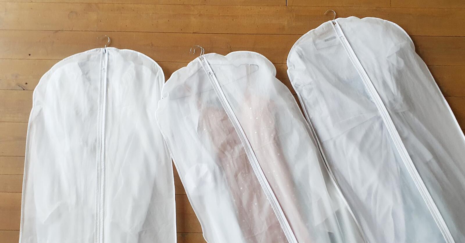 Three white hanging garment bags with garments inside of them are laying on a warm-toned wood floor.