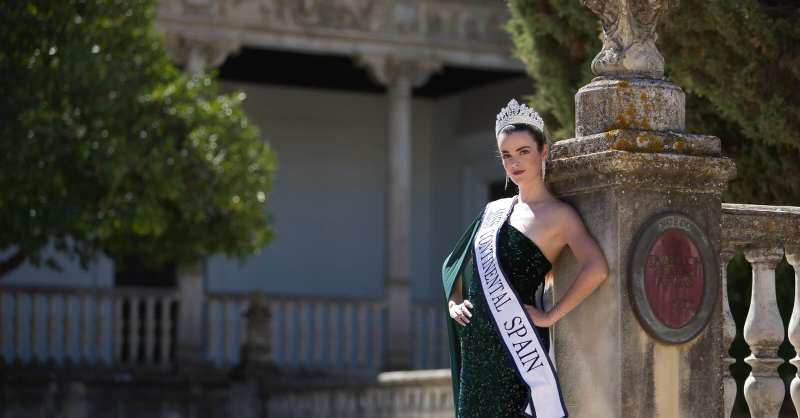 A woman leaning against a stone column. She is wearing a green sequined dress, a crown, and a “Miss Continental Spain” sash.