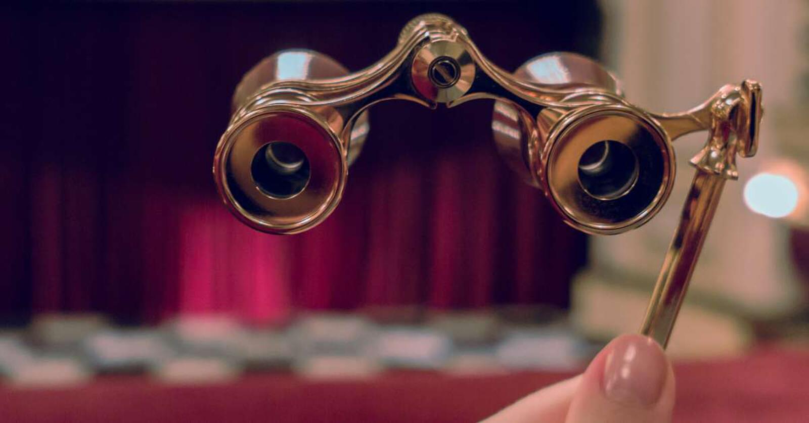 A hand holding up a pair of gold opera glasses. In the blurred background are black and white tiles and a red stage curtain.