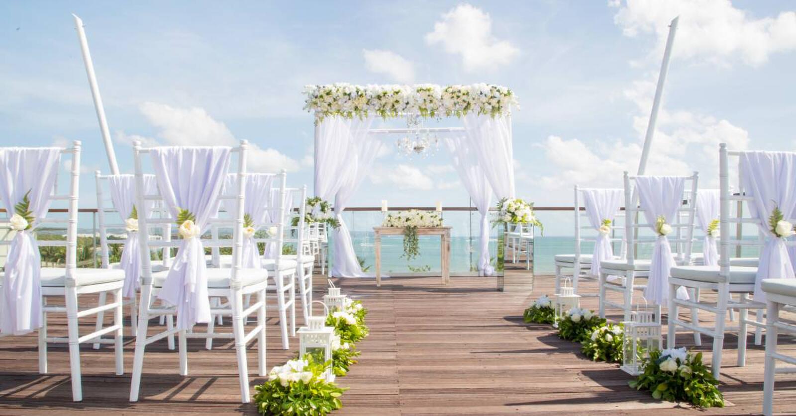 Rows of decorated white chairs facing a wedding arch with white curtains and flowers. The ocean is visible beyond the arch.