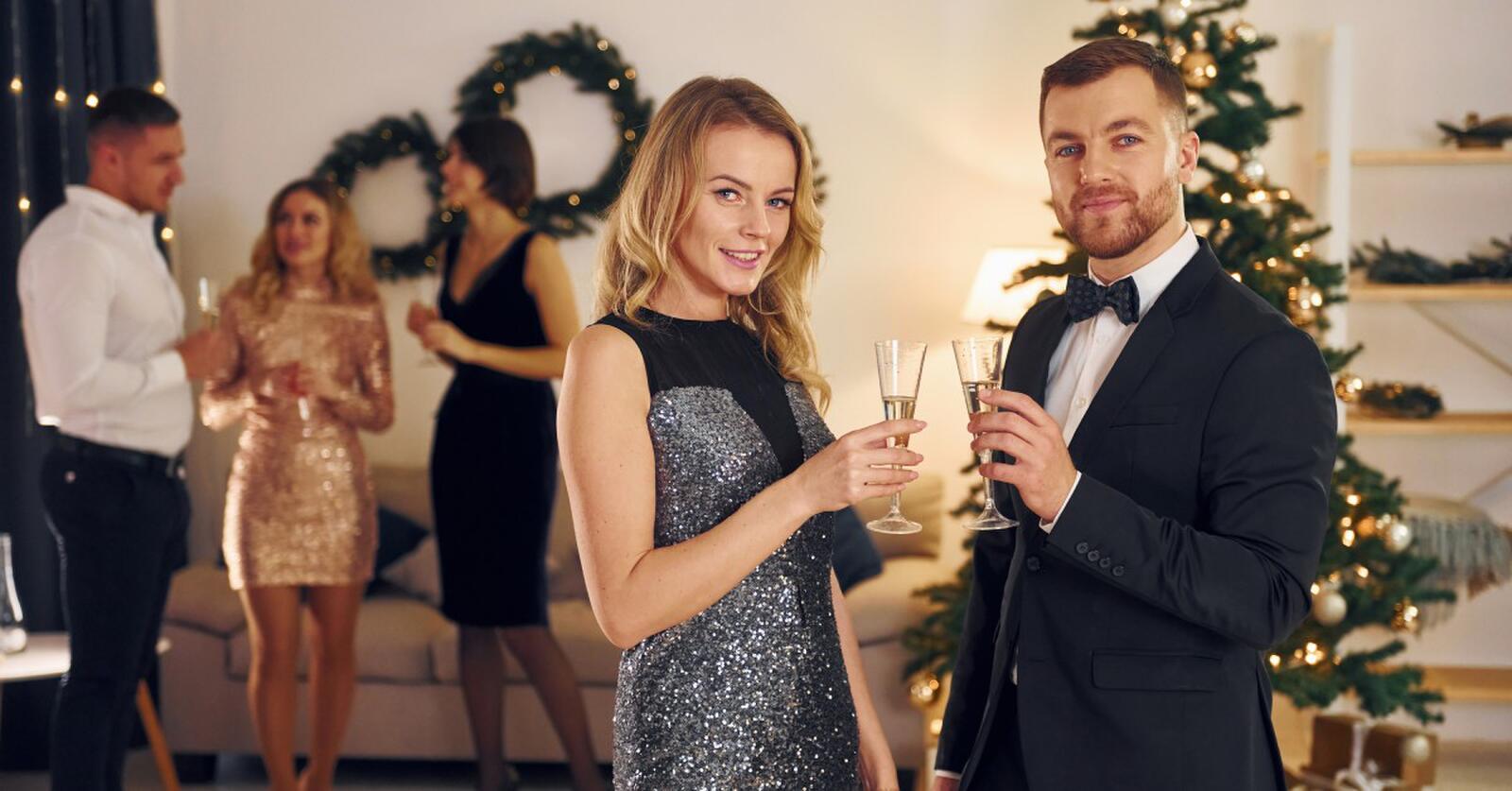 A man in a suit and a woman in a sparkly silver dress smile and pose with champagne in front of a Christmas tree.