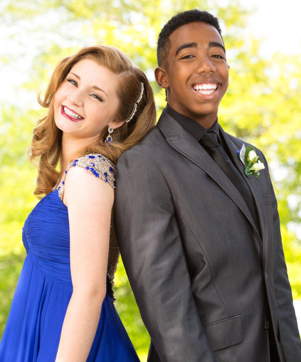 A teen girl in a blue prom dress and a teen boy in a gray suit smiling at the camera as they stand back to back.