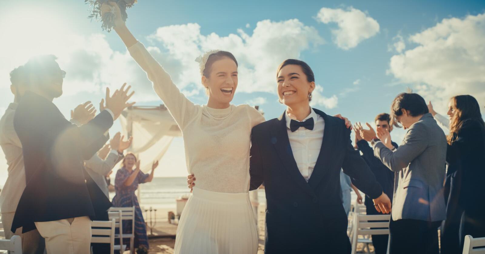 Two women smile as they walk away from a wedding arch on a beach. The guests around them clap.