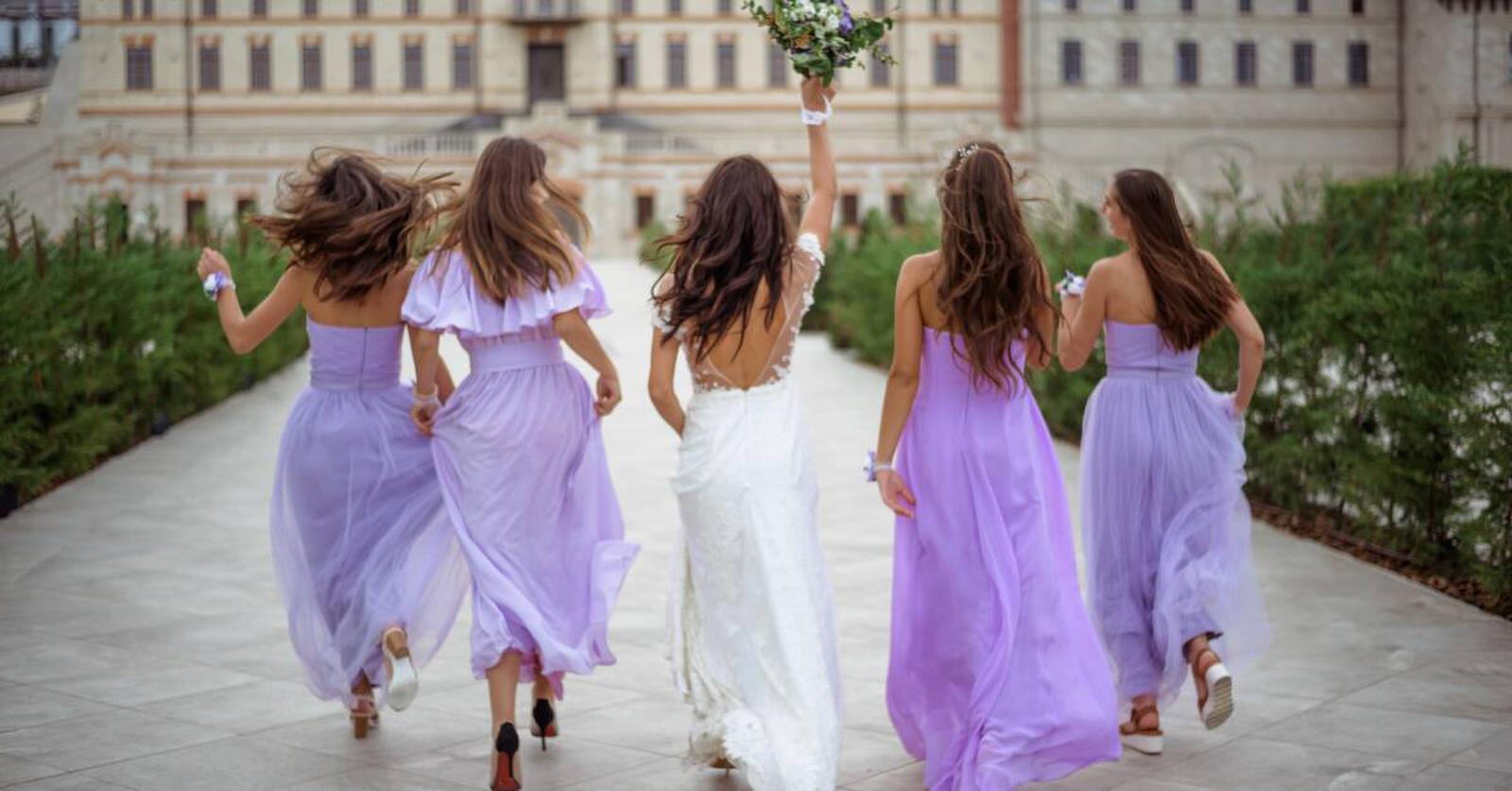 A bride and four bridesmaids in purple dresses run away from the camera toward a large building. The bride is holding a bouquet.