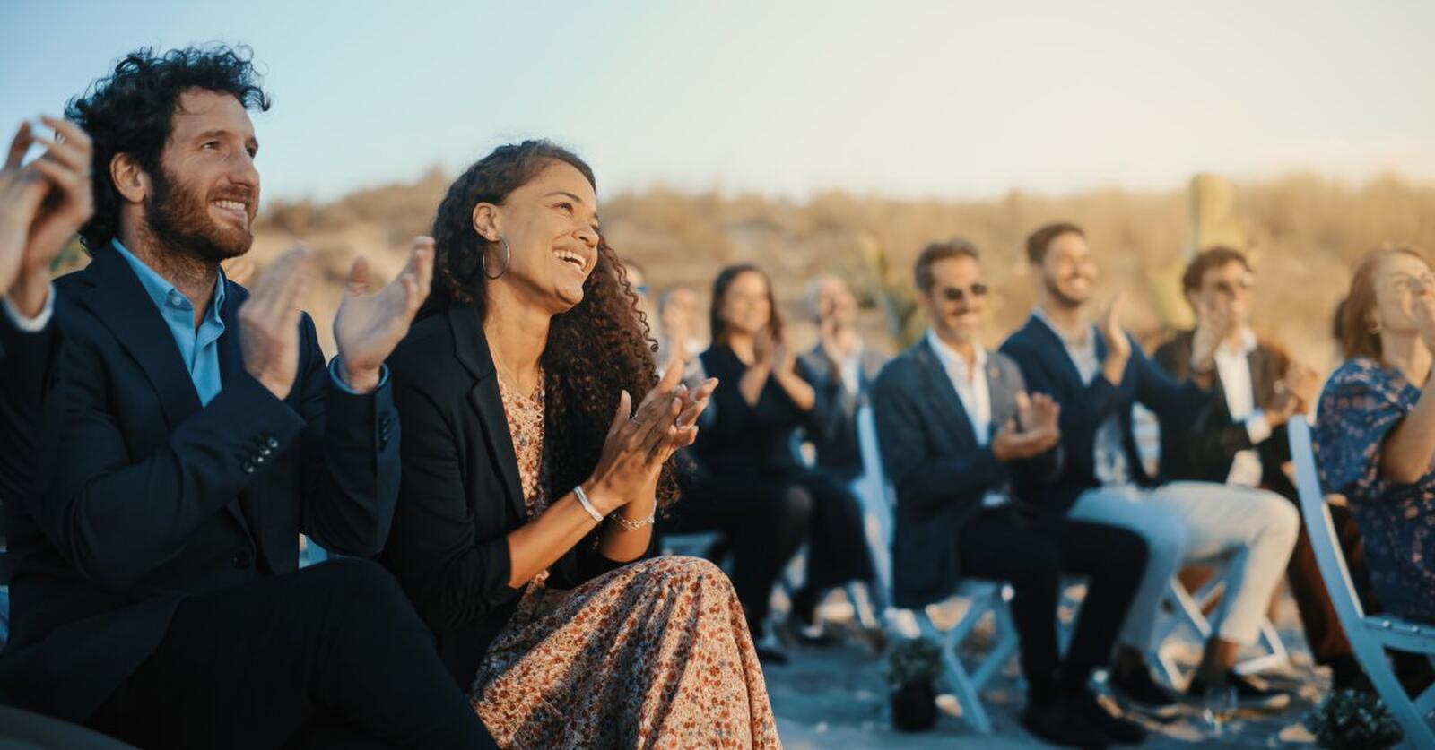 A group of people in suits and dresses smiling and clapping while sitting in white folding chairs outside.